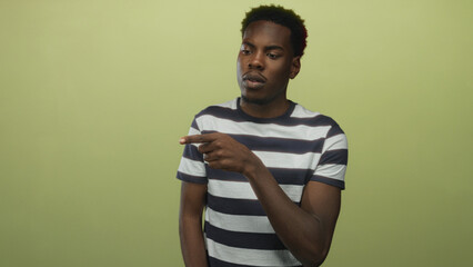 Young african american man wearing striped shirt pointing finger at unseen object in green studio;...
