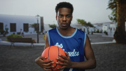 Fototapeta premium Man wearing blue basketball jersey holds basketball on sunny street beside wooden bench; confidence aspiration.