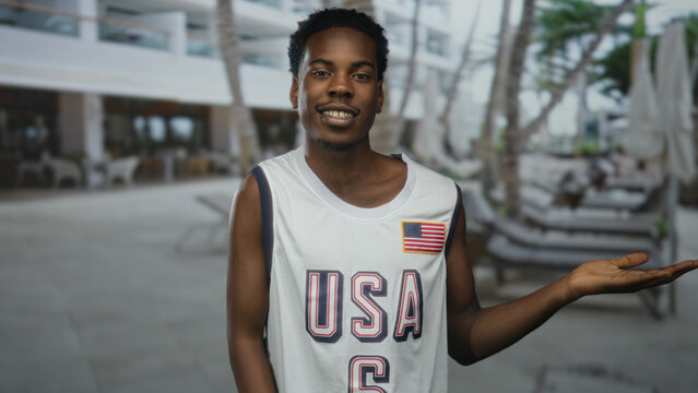 Young african american man wearing white basketball jersey with usa flag patch holds open palm gesture at building; confidence.