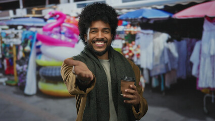 Smiling man in market street holding coffee with vibrant stalls in background reaching playfully forward.