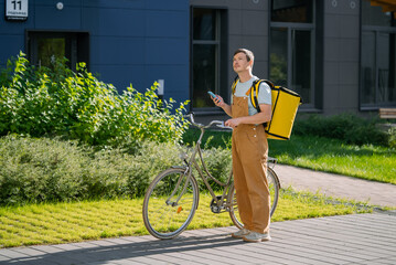 Male bike courier in overalls with a yellow delivery backpack checks smartphone while standing with his bicycle on a busy city sidewalk, ready to deliver parcels or food