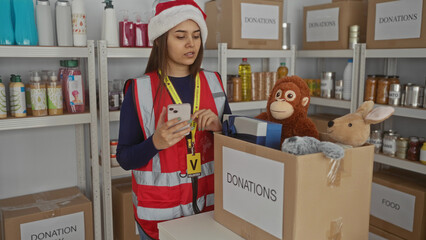 Woman in santa hat using smartphone at charity center surrounded by donation boxes and toys, indoors during christmas, showcasing volunteering dedication for community support.