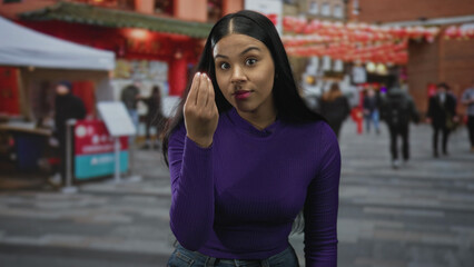 Fototapeta premium Young latin woman making pinched fingers gesture on crowded urban street lined with market stalls; confusion.