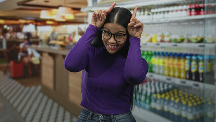 Fototapeta premium Woman showcasing hands in rock horns gesture while standing by shelves at restaurant counter; playfulness.