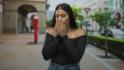 Latin woman hugging arms on street pavement lined with arches and lampposts in daylight;...