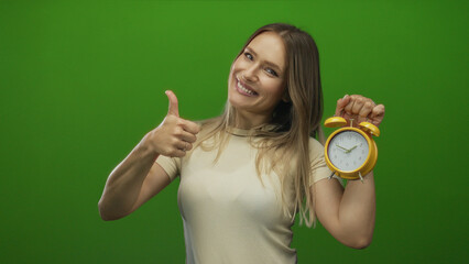 Young woman with blonde hair holding a yellow alarm clock smiling over green isolated background while giving a thumbs up