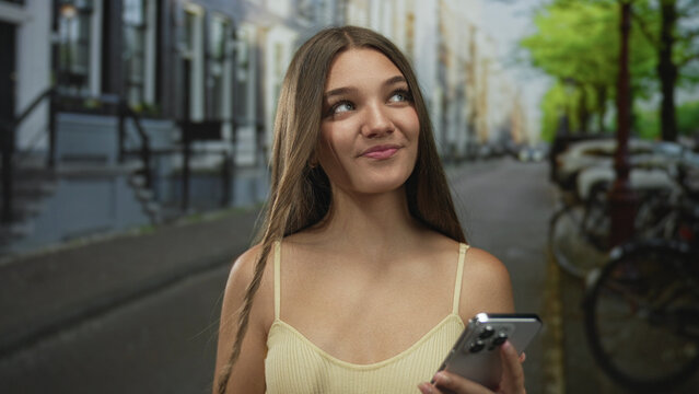 Teenage girl smiles and holds smartphone on busy street; digital social connection happiness. - Powered by Adobe