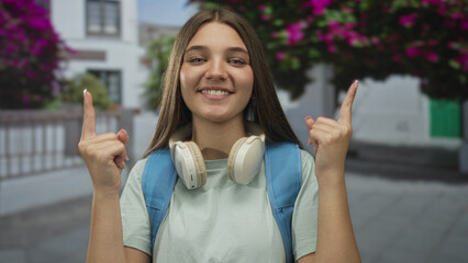 Woman wearing backpack and headphones points finger upward in sunlit street lined with...