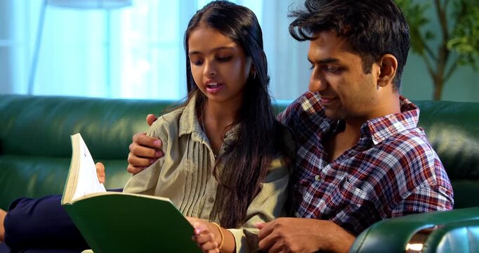 Reading book together, Indian father and daughter sitting on sofa in modern home, Asian dad bonding with young girl while sharing story, family quality time, indoor learning and emotional connection