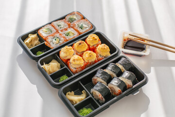 Assorted sushi rolls in black takeout boxes with soy sauce, chopsticks, and ginger on a white table, symbolizing Japanese cuisine, food delivery, and modern dining culture