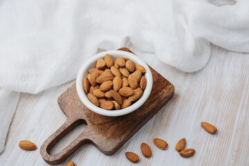 Whole almonds in bowl on wooden board, scattered nuts on light surface, natural food.