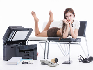 Asian pretty young woman confident braided hair in office costume lying on bench, poses playfully beside large printer books sunglasses keyboard landline phone shoes in white studio fashion setting.
