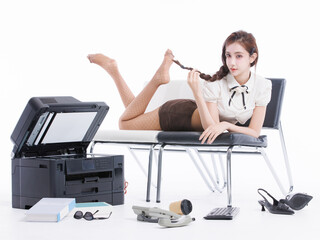 Asian pretty young woman confident braided hair in office costume lying on bench, poses playfully beside large printer books sunglasses keyboard landline phone shoes in white studio fashion setting.