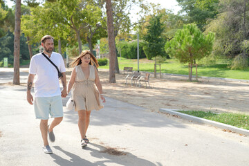 Fototapeta premium A man and woman are walking together in a park. The man is wearing a white shirt and blue shorts, while the woman is wearing a dress. They are holding hands and walking down a path