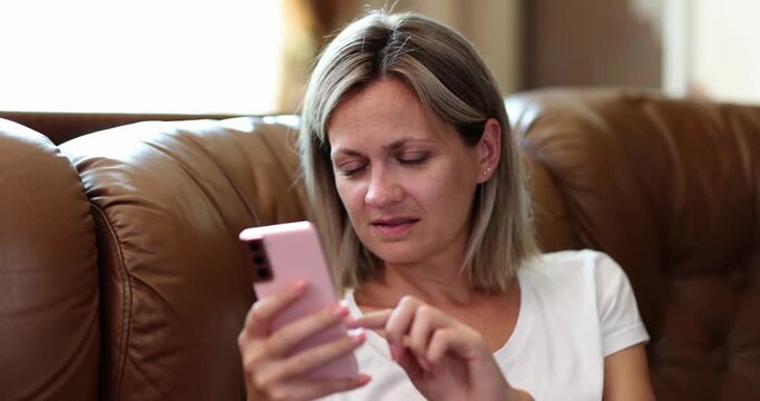 Adult woman sits on leather couch with smartphone frowning at screen. User taps display checking message content wasting time during break slow motion