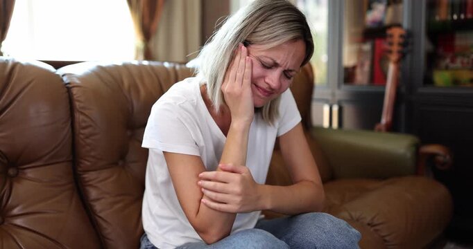 Sad woman suffers from strong toothache sitting on leather sofa in living room. Blonde female holds cheek with hand trying to reduce jaw pain at home