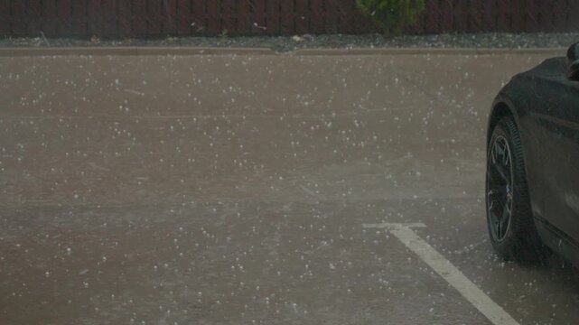 Torrential rain and hail fall on a city parking lot, creating a rapid buildup of water over the asphalt. The storm intensifies, impacting visibility and surroundings.