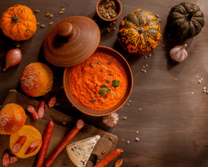 Homemade creamy pumpkin soup garnished with seeds, bakery buns, blue cheese, spicy sausage, and colorful autumn gourds on dark rustic table, cozy fall comfort food, warm festive kitchen mood