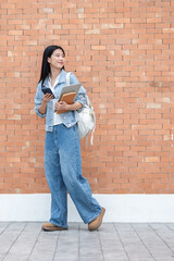 Asian woman student holding phone with laptop on book turning head back as she walking by brick wall