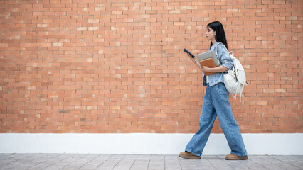 Asian woman student looking at phone holding laptop and book while walking in front of a brick wall.