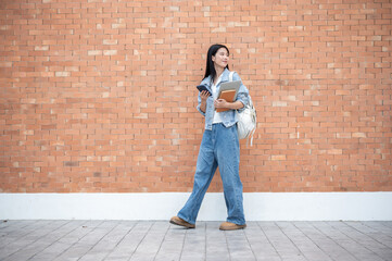 Asian woman student holding phone with laptop on book turning head back while walking by brick wall.