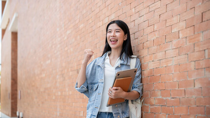 Happy asian woman student holding laptop and book raising a fist standing leaning against brick wall