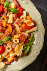 Close-up italian pasta salad with diced cheese, salami, cherry tomatoes, olives and fresh basil on ornate ceramic plate, vibrant gourmet Mediterranean lunch, healthy festive food, natural daylight