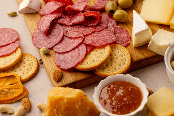 close-up charcuterie board with thinly sliced salami, round crackers, brie wedges, hard cheese, green olives, nuts and sweet fruit preserves on wood, festive gourmet party snack in daylight