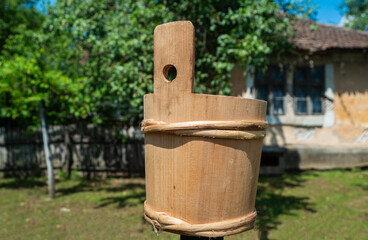 Rustic wooden bucket, handmade in traditional style, placed outdoors in a countryside setting with an old house and garden in the background. Traditional handmade container symbolizing rural life