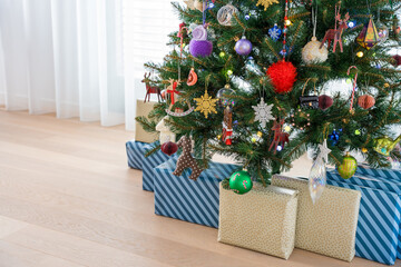 Beautifully wrapped presents in blue and gold paper placed under an artificial Christmas tree adorned with colorful ornaments and glowing lights, creating a warm festive holiday atmosphere at home