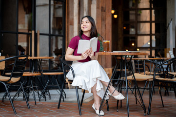 Fototapeta premium Asian woman holding book aside coffee and laptop while sitting cross legged at wooden table in cafe.