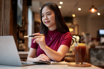 Asian woman holding pen pointing and looking at a laptop aside coffee while sitting at table in cafe