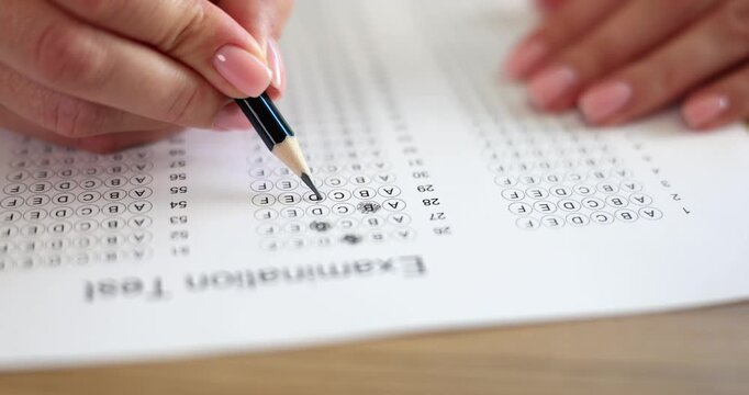 Woman student fills out multiple choice exam answer sheet with pencil on desk. Female hand chooses bubble with letters for options in classroom