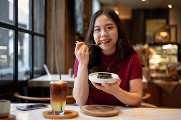 Young asian woman smiling and holding spoon enjoy eating chocolate cake aside orange coffee in cafe.