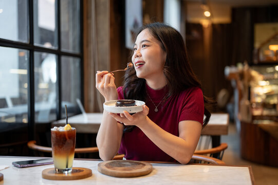 Smiling young asian woman holding spoon and plate enjoy eating chocolate cake at table in cafe.