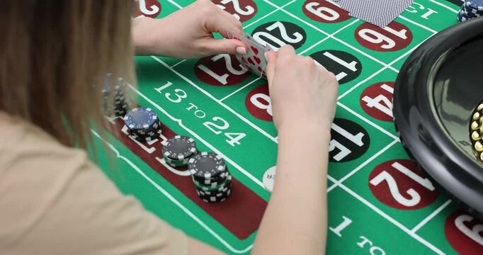 Player sits at roulette table holding cards near chips and wheel. Gambler checks hand and adjusts bet covering numbers while waiting for next spin
