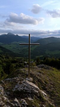 Vertical Cross on rocky hilltop overlooking green valley with forests and mountains in background