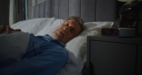 Fototapeta premium Mature Man Slowly Wakes Up Early In Morning In Bedroom Filled With Bright Sunlight. Digital Alarm Clock Shows 7 AM. Slow Morning, Time in Bed. Concept of Morning Routine, Good Morning. Static Shot.