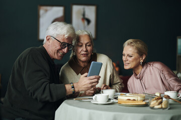 Senior Caucasian man showing smartphone to elderly female friends while sitting together at table with coffee and cake, all smiling and looking at device during social reunion