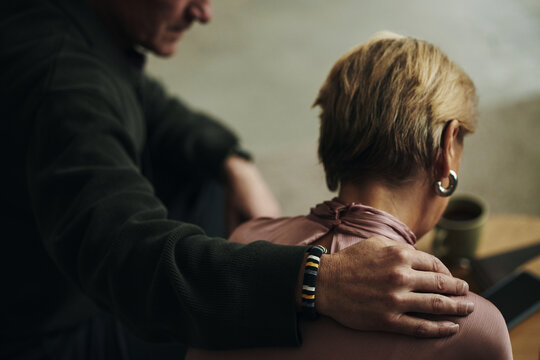 Senior Caucasian man comforting wife or female friend or relative by placing hand on her shoulder, both sitting closely together, woman facing away, during grief or bereavement