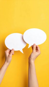 Pair of hands holding white paper speech bubbles against a vibrant yellow background, symbolizing communication and dialogue