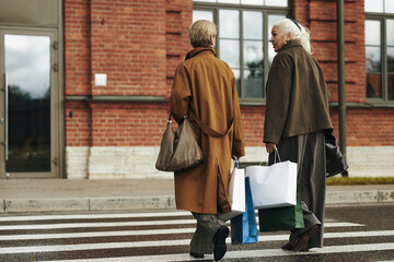 Naklejka premium Two senior Caucasian women walking across crosswalk carrying shopping bags and handbags, engaging in conversation, urban background with brick building visible behind them