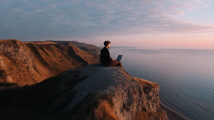 Man sitting on the edge of a cliff overlooking the ocean. he is wearing a black jacket and pants and is holding a laptop in his lap.