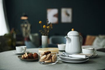 Table set with glass bowl of assorted cookies, plates with napkins, cups, and ceramic teapot arranged for serving snacks and hot beverages in cozy indoor setting