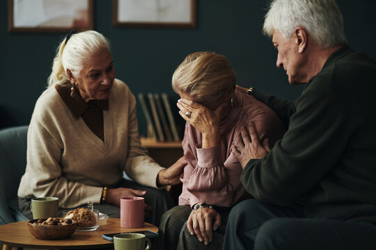 Three senior Caucasian people comforting each other, two women and one man sitting together, one woman covering face with hand while others offering support and empathy in bereavement or grief