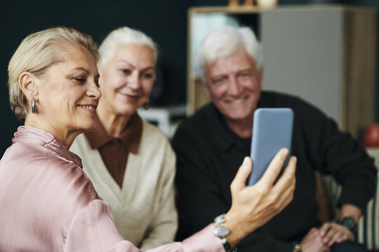 Senior Caucasian woman taking selfie with smartphone while sitting with male and female friend couple, all smiling and looking at device together while taking photo for social media