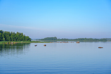 Calm lake with forested shoreline and scattered rocks under blue sky