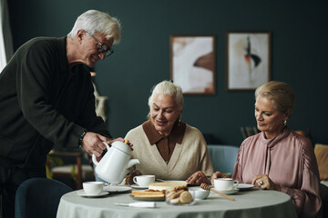 Senior Caucasian man pouring tea for two elderly women sitting at round table enjoying conversation and desserts in cozy indoor setting, all friends smiling and engaged