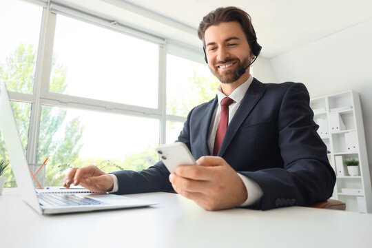 Young male secretary with headset, mobile phone and laptop working at table in office