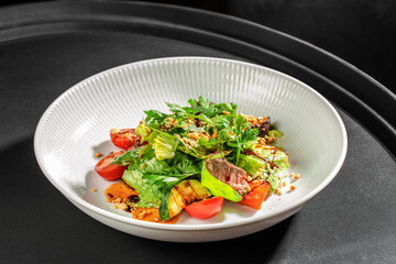 Fresh mixed salad with grilled vegetables, beef slices, and crunchy toppings served in a white bowl on a dark background.